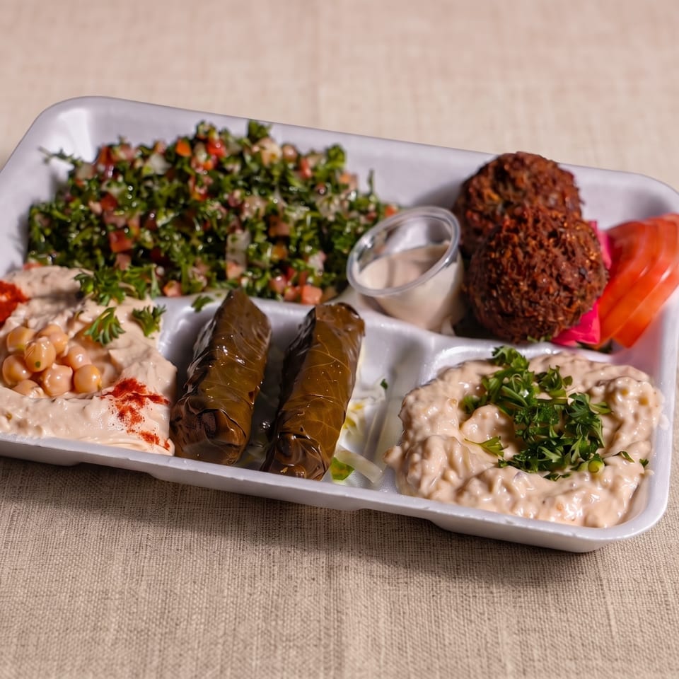 Mediterranean platter featuring falafel, stuffed grape leaves, tabbouleh, hummus, creamy salads, tahini, and sliced tomatoes.